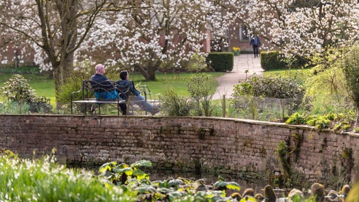A man and a woman sat on a bench facing away from the camera. They are sat underneath white blossom trees. The red brick Georgian house of Dunham Massey can be seen in the background. A brick canal wall surrounded by green flowers can be seen in the foreground.